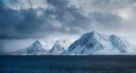 Majestic snow-capped mountains rise from the deep blue ocean under dramatic cloudy sky. Winter landscape with snowy rocks, overcast sky and sea in the Lofoten islands, Norway. Nature. Arctic scenery