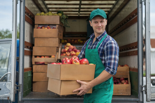 Confident delivery worker carrying fresh apples in a cardboard box from a truck for organic farming and healthy food lifestyle promotion