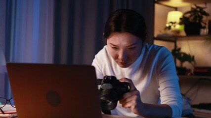 Portrait of pretty young asian woman photographer hold digital camera looking at screen choosing photos for editing while sitting in front of laptop computer at home workplace at late night