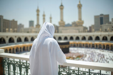 A Muslim woman in white abaya and hijab on a balcony of the Great Mosque of Mecca looking at the procession of faithful pilgrims circling the Kaaba performing the Hajj, fulfilling a pillar of Islam.