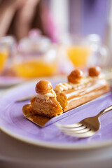Caramel eclair lies on a purple plate with a gold fork on a white table in a coffee shop.