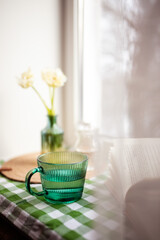 A green glass cup of water stands on the windowsill. White tulips are in a vase, and an open book lies next to it. Winter window view.
