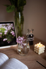 A branch of lilac in a glass of water. A white square bubble candle is lit. A book is on the wooden table.