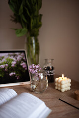 A branch of lilac in a glass of water. A white square bubble candle is lit. A book is on the wooden table.