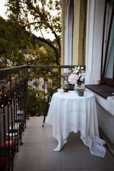Bouquet of ranunculus blooms in a glass vase standing on a white table with cup of tea on a french balcony in the evening with street lights on its background in spring