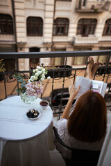 Woman reading a book. Bouquet of ranunculus blooms in a vase standing on a white table with cup of tea on a french balcony in the evening with street lights on its background