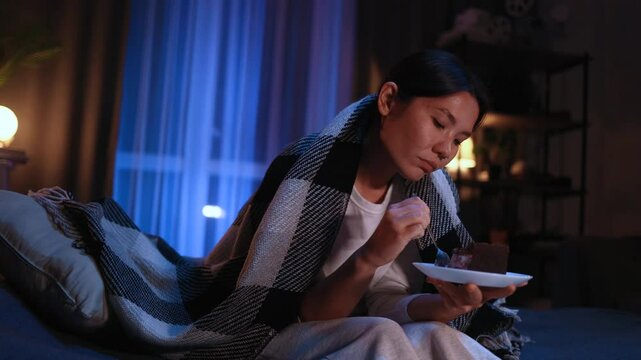 Sad young asian woman dealing with stress by eating food sitting on bed late at home Upset female wrapped in blanket crying and eating cake at night indoors alone Mental heath problem