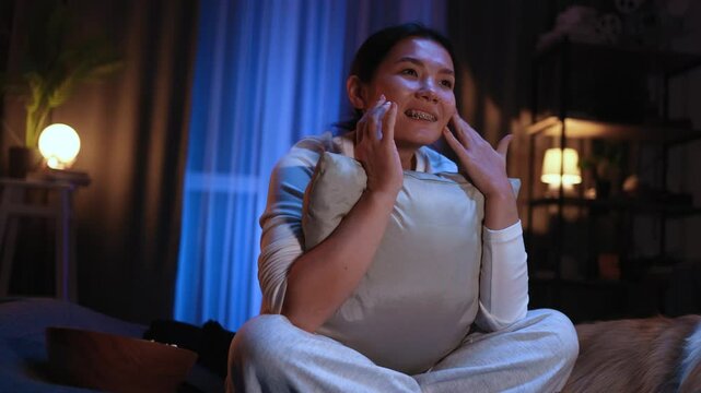 Portrait of sensitive young asian woman hugging pillow sitting on bed and watch drama film series at home indoors Beautiful sad female wipes away tears after watching love soap movie alone
