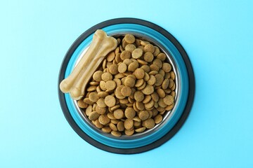 Dry pet food in feeding bowl and chew bone on light blue background, top view