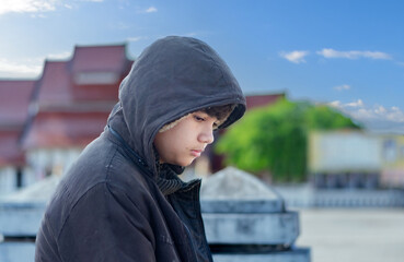 Asian young boy wearing a dark hooded jacket, sitting near a stone fence or barrier thoughtfully...