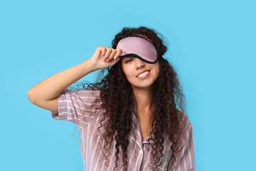 Beautiful young happy African-American woman in pajamas with sleep mask on blue background