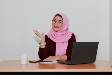 An Asian Muslim lady smiling while pointing with palm of her hand to presenting or explaining something.