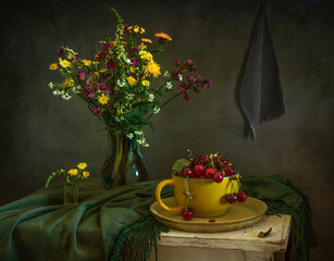 Still life with ripe cherries in a large yellow cup and a bouquet of meadow flowers.