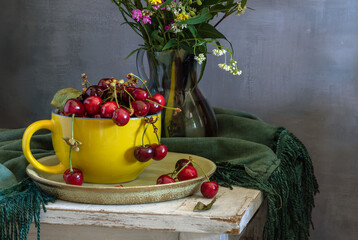 Still life with ripe cherries in a large yellow cup and a bouquet of meadow flowers.