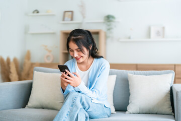 Smiling pretty young Asian woman sitting on cozy couch, using funny mobile apps in living room. Woman at Home, Doing Online Shopping, Messaging Friends, Posting on Social Media © Monster Ztudio