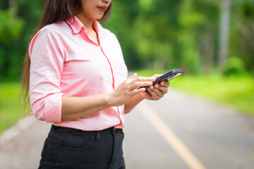 A young woman walks along a grassy path in a green forest, enjoying sunlight and nature. She relaxes, looks at her new mobile phone, and embraces the peaceful outdoor environment