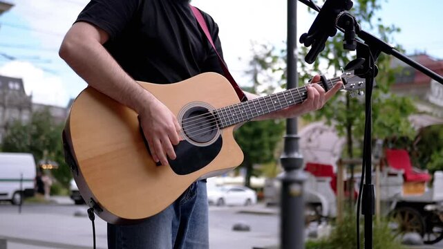 Close-up of a guitarist's hands skillfully playing an acoustic guitar on a city street. The blurred background hints at the bustling urban life surrounding the performance.