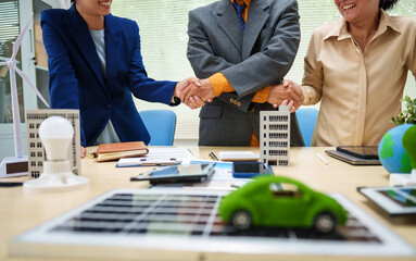 Asian business professionals in collaborate at desk with building models, planning sustainable projects solar panels, electric cars, wind turbines, eco-friendly clean energy, shaking hands