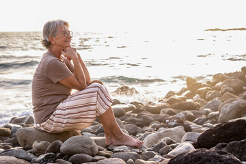 Portrait of attractive relaxed senior woman barefoot sitting at the beach at sunset looking away smiling - elderly lady enjoys vacation and freedom