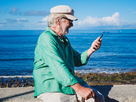 Smiling senior retired man in white hat sitting near the beach using mobile phone looking at social - retired life, horizon over the sea