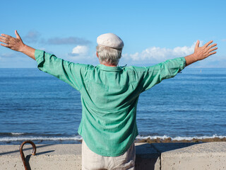 Rear view of caucasian senior retired man standing face the sea with outstretched arms enjoying freedom and a sunny day. Authentic retirement lifestyle