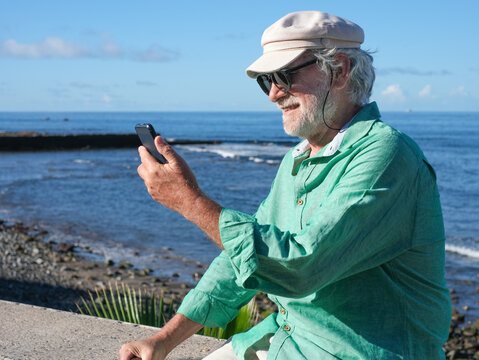 Portrait of smiling senior retired man in white hat sitting near the beach using mobile phone looking at social - retired life, horizon over the sea - Powered by Adobe