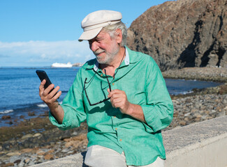 Portrait of smiling senior retired man in white hat sitting near the beach using mobile phone looking at social - retired life, horizon over the sea