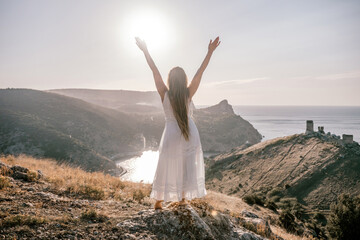 A woman stands on a hill overlooking the ocean. She is wearing a white dress and has her hands on...