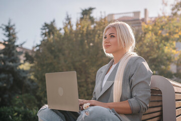 A woman is sitting on a bench with a laptop in front of her. She is smiling and she is enjoying her time.