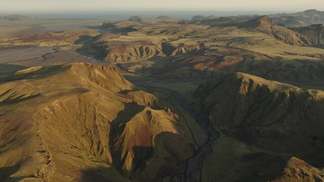 A wide aerial view of Thakgil, Iceland, revealing sprawling volcanic highlands, winding valleys, and streams under soft golden sunlight.