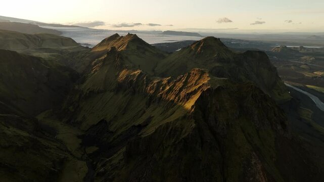 A stunning aerial view of Thakgil, Iceland, highlighting sunlit volcanic peaks, deep valleys, and distant rivers under a serene sky.