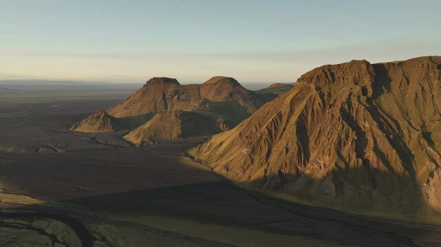 Aerial view of majestic, sunlit mountains in Thakgil, Iceland, showcasing rugged cliffs, expansive plains, and the serene beauty of golden hour.