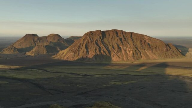 Aerial view of sunlit mountains in Thakgil, Iceland, casting long shadows over the expansive plains, showcasing rugged and dramatic terrain.