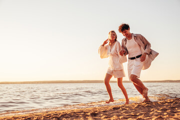 Smiling beautiful woman and her handsome boyfriend. Excited couple in casual clothes. Happy cheerful family. Female and man run at sunrise over sea beach outdoors. Seaside in summer day