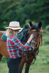 Obraz premium Happy blonde with horse in forest. Woman and a horse walking through the field during the day. Dressed in a plaid shirt and black leggings.