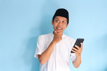 Young asian muslim man smiling holding his mobile phone while looking side with touching his chin