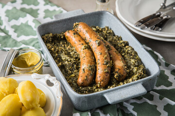 Oldenburger Grünkohl mit Pinkel Kohlwurst in Schale, Salzkartoffeln und Senf auf gedeckten grau Tisch mit grün Tuch, Teller, Besteck rustikal 