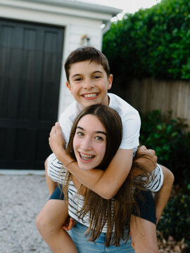 Teenage girl giving her brother a piggyback around their yard at home
