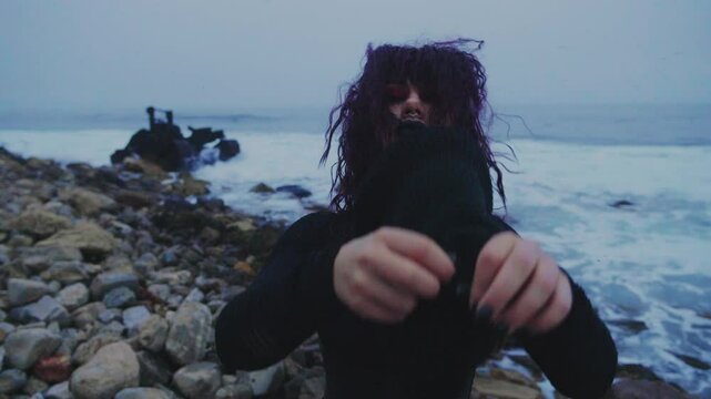 A masked figure in black stands at the seashore and pulls off a torn black mask, revealing a young Goth girl. Mysterious masked woman at the rocky seacoast.
