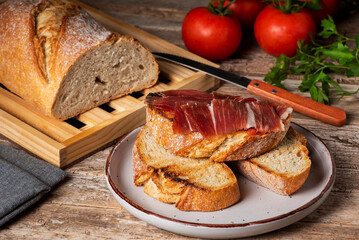 Several slices of bread, one of them with tomato and ham, next to a loaf of bread and some tomatoes, on a rustic wooden background.