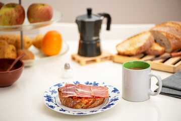 Serrano ham toast, olive oil and tomato next to a cup of coffee with milk, on the kitchen table and next to more slices of toast, a coffee maker and a fruit bowl.