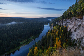 A tranquil river flows gently through lush green forests and towering cliffs at dusk, with mist rising above the water and vibrant autumn foliage illuminating the scene