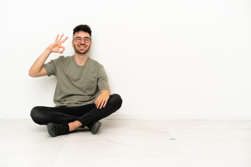 Young caucasian man sitting on the floor isolated on white background showing ok sign with fingers