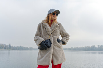 A girl person stands near a misty lake, donning a light fur coat, black gloves, red pants, and a gray cap, holding a black handbag with a gold chain strap.