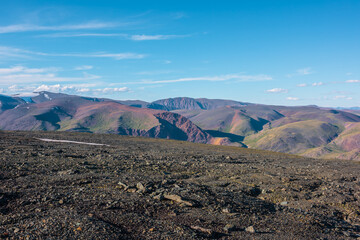 Scenic top view from sunlit stony plain to big colorful mountain range in bright sun under clouds in blue sky. Layered alpine scenery with large multicolor mountains of many vivid colors in sunny day.
