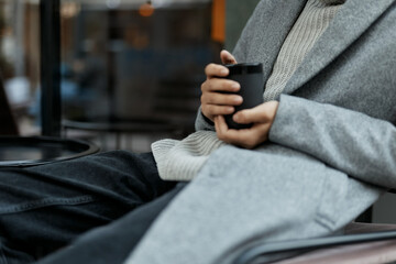 Stylish man in gray jacket and black pants sitting in chair with cup of coffee in hand