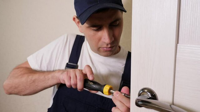 Installation of a lock on the front wooden entrance door. Portrait of young locksmith workman in blue uniform installing door knob. Professional repair service. Maintenance Concept.