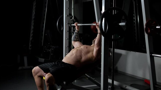A man with a naked torso does bench press in a dark gym. A man lifts a barbell in a Smith simulator. A male bodybuilder trains in the gym, shadows beautifully emphasize a muscular body. Lifts weights.