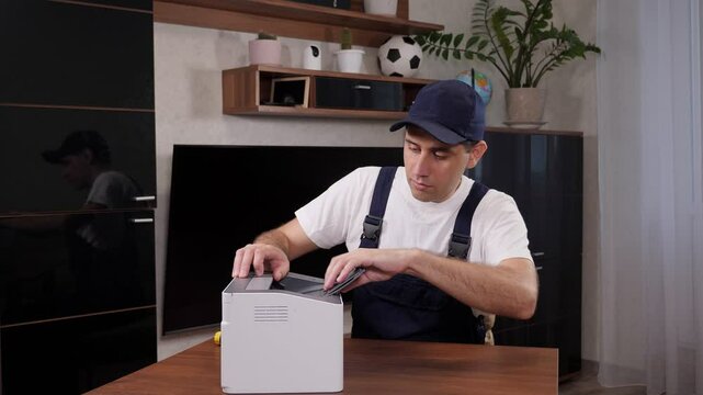 Close-up of a handyman in overalls, a white t-shirt and a cap with a screwdriver in his hands repairs the printer in the office or at the client's home. Call a home repairman.