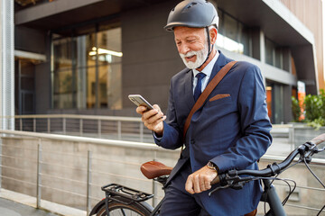 Old bearded businessman with helmet and bicycle texting over smartphone while commuting to work in...